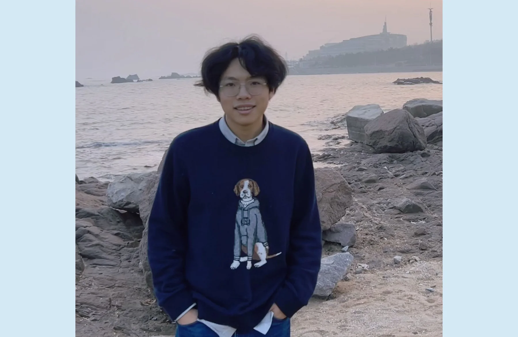 A picture of Patton Yin standing on the beach in front of the sea with sweatshirt showing a dog.