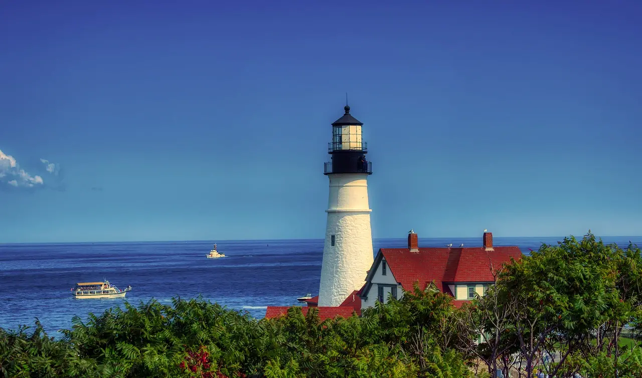 A photo of the Portland Head Lighthouse looking from to the sea with boats to its left.